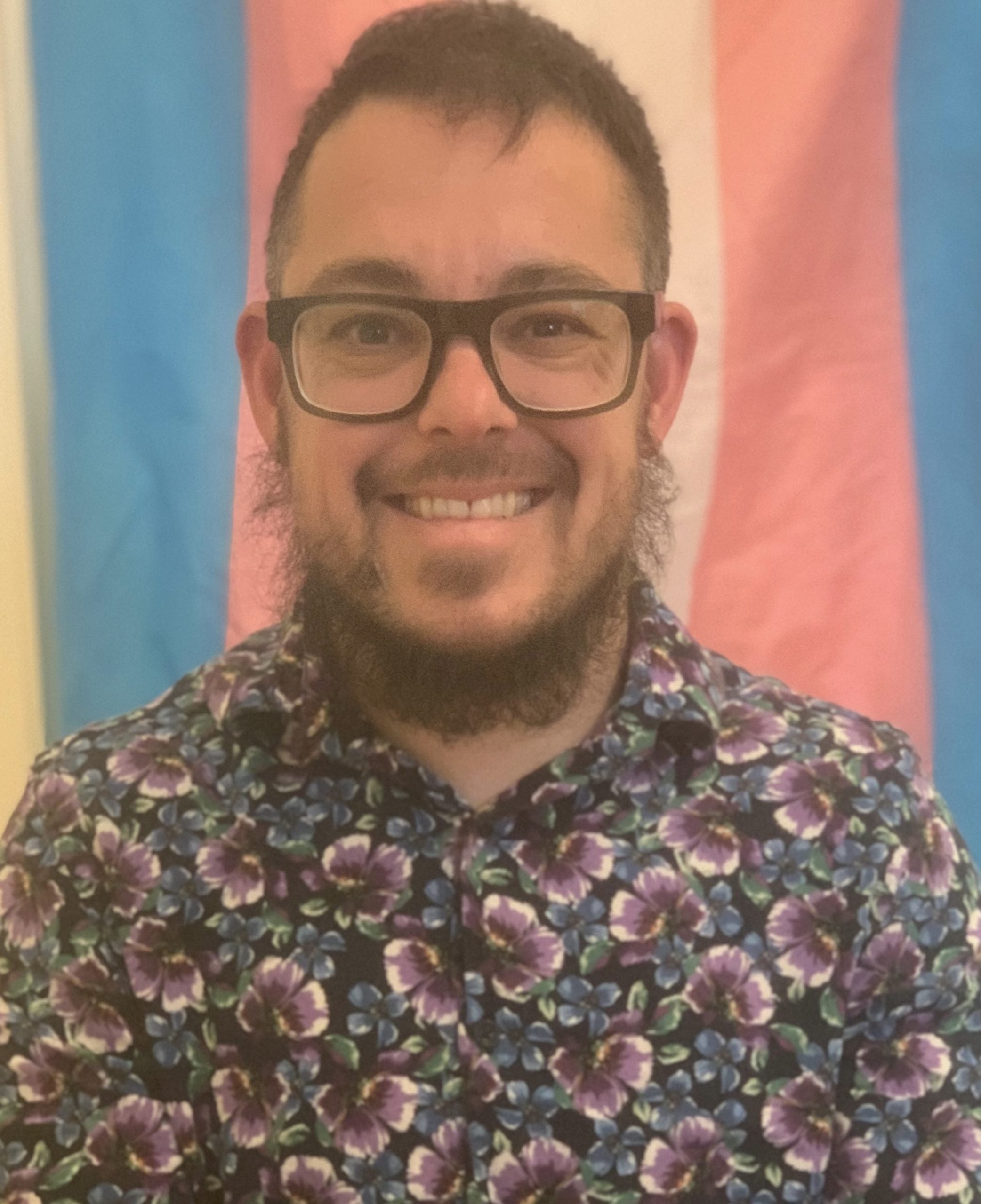 Jax is a caucasian person with glasses, short hair and a beard. He is wearing a flowery top